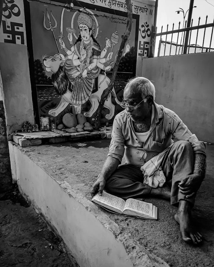 An elderly man reads a book near a vibrant Hindu temple painting.