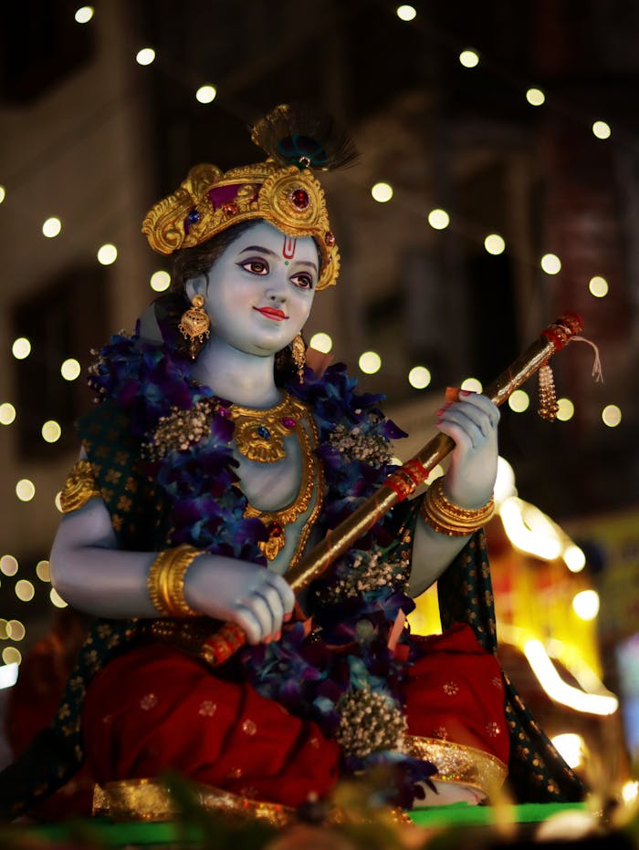 Colorful Krishna statue adorned with flowers and lights during a Hindu festival at night outdoors.